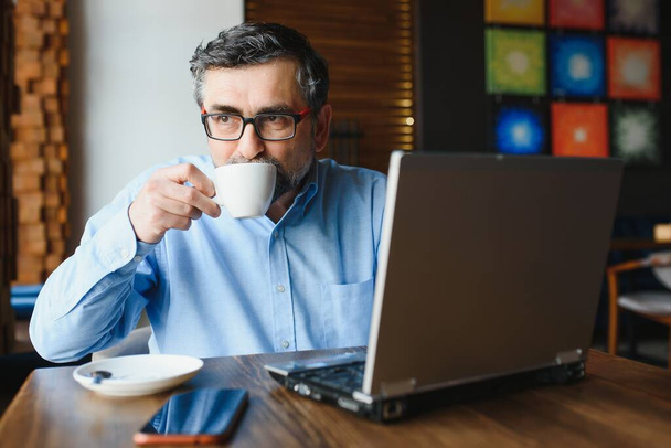 Male freelancer is working in a cafe on a new business project. Sits at a large window at the table. Looks at a laptop screen with a cup of coffee. - Foto, afbeelding