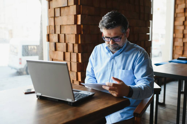 Άνδρας ελεύθερος επαγγελματίας εργάζεται σε ένα καφέ σε ένα νέο επιχειρηματικό σχέδιο. Κάθεται σε ένα μεγάλο παράθυρο στο τραπέζι. Κοιτάζει μια οθόνη laptop με ένα φλιτζάνι καφέ. - Φωτογραφία, εικόνα