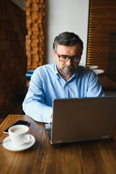 Άνδρας ελεύθερος επαγγελματίας εργάζεται σε ένα καφέ σε ένα νέο επιχειρηματικό σχέδιο. Κάθεται σε ένα μεγάλο παράθυρο στο τραπέζι. Κοιτάζει μια οθόνη laptop με ένα φλιτζάνι καφέ. - Φωτογραφία, εικόνα