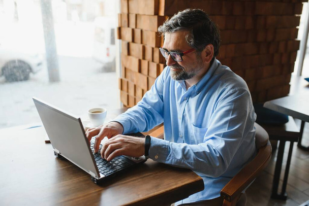 Mature businessman drinking coffee in cafe. Portrait of handsome man wearing stylish eyeglasses using laptop, looking at camera, smiling. Coffee break concept. - Foto, Bild