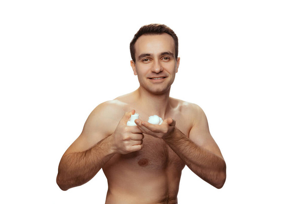 Portret van een jonge, shirtloze man die scheerschuim op de hand aanbrengt, geïsoleerd over witte studioachtergrond - Foto, afbeelding