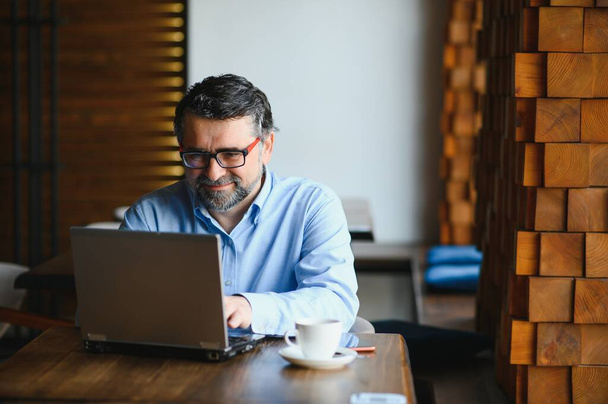 Mature businessman drinking coffee in cafe. Portrait of handsome man wearing stylish eyeglasses using laptop, looking at camera, smiling. Coffee break concept. - Photo, Image