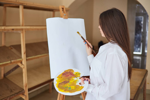 白いシャツを着た若いブルネットのアーティストが彼女のスタジオでキャンバスにペイントします。彼女の隣にはイーゼル、絵画、様々な芸術技術があります。. - 写真・画像
