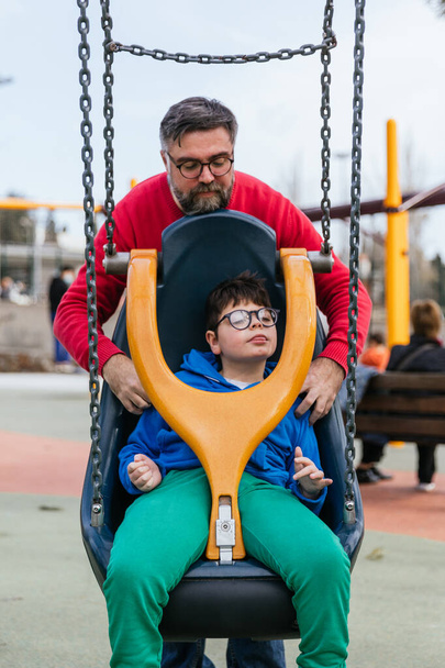 Een ontspannen kind met een handicap spelen op aangepaste schommel met zijn vader in een speeltuin - Foto, afbeelding