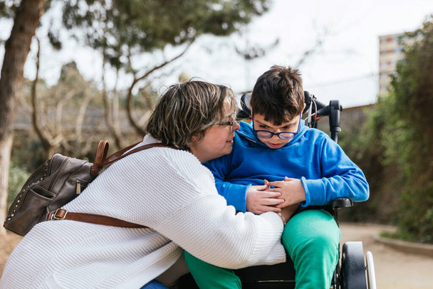 Kochająca matka przytulająca syna niepełnosprawnego na wózku inwalidzkim w parku. - Zdjęcie, obraz