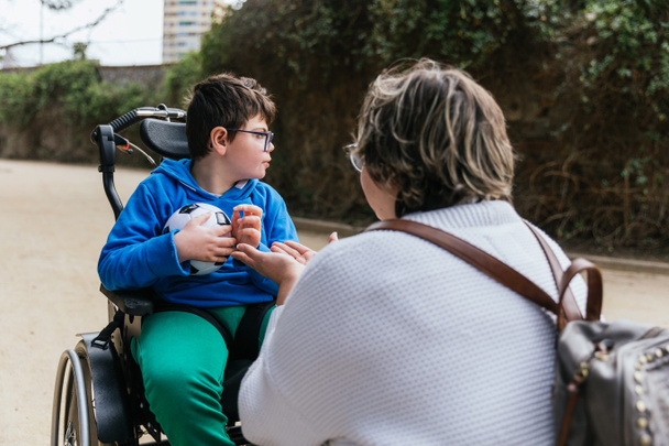 Een kind met een handicap in een rolstoel met een voetbal waarmee hij met zijn moeder in het park speelt - Foto, afbeelding