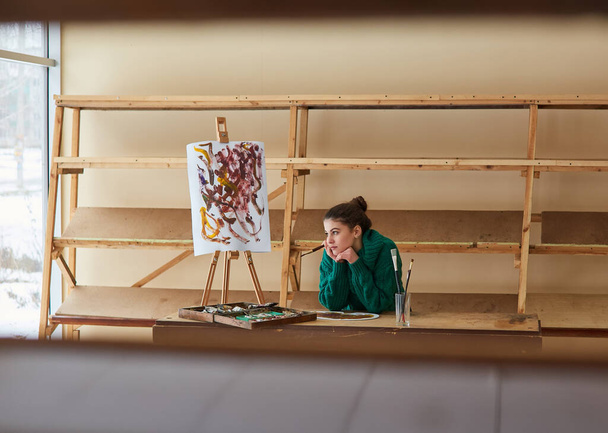 brunetka artystka dziewczyna stoi w pobliżu sztalugi i hoslt z pędzlami w rękach. Kreatywne otoczenie w pracowni sztuki - Zdjęcie, obraz