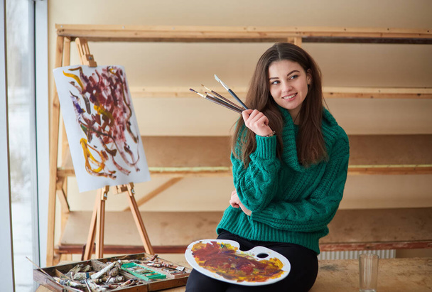 brunette artist girl stands near the easel and hoslt with brushes in her hands. Creative setting in the art workshop - Photo, Image