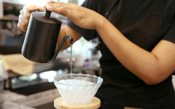 Μια γυναίκα καφετζού φορώντας ποδιά ρίχνει ζεστό νερό πάνω από καβουρδισμένους κόκκους καφέ για να προετοιμάσει καφέ για τους πελάτες στο κατάστημα. - Φωτογραφία, εικόνα