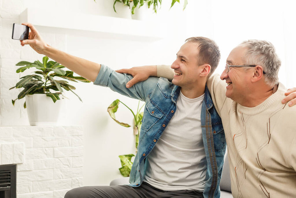 Glücklicher Moment. fröhlicher junger Mann, der ein Selfie mit seinem fröhlichen älteren Vater macht, der freundlich in die Kamera winkt und lächelt - Foto, Bild