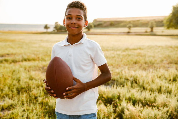 Black boy wearing white shirt smiling while posing with rugby ball outdoors - Valokuva, kuva