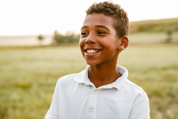 Black boy wearing white shirt smiling and looking aside while posing outdoors - Fotó, kép