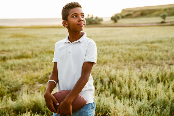 Black boy wearing white shirt posing with rugby ball outdoors - Фото, изображение