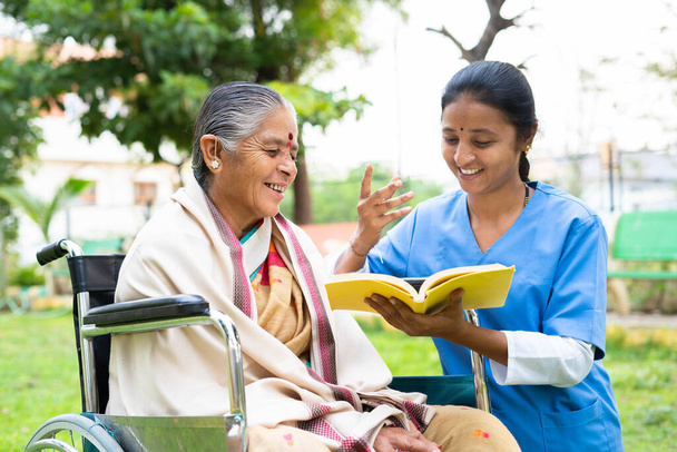 Sairaanhoitaja ja vanhempi nainen pyörätuolissa nauramassa lukemalla kirjaa tai romaania puistossa - käsite rentoutuminen, ammatillinen ammatti, terapia tai hoito - Valokuva, kuva