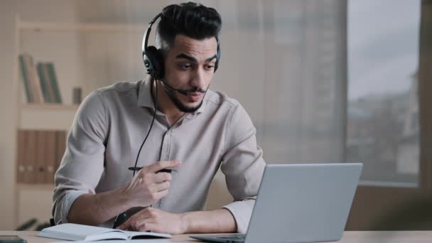 Focused man hispanic businessman interpreter teacher student wear headset watching online webinar on laptop holding video call conference making notes writing at work desk learning education course - 映像、動画