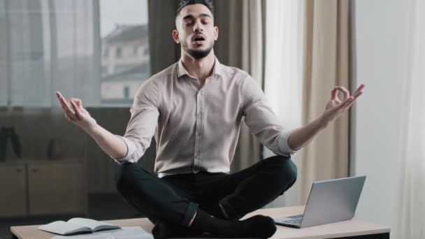 Mindful calmo arabo uomo d'affari dipendente in posizione di loto sedersi sulla scrivania di lavoro occhi chiusi meditando prendere pausa a casa ufficio mantenere equilibrio mentale pazienza yoga pratiche senza stress sentire zen equilibrio - Filmati, video