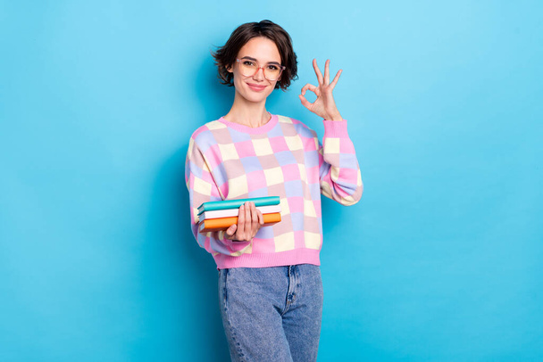Photo of cute millennial brunette lady hold book show okey wear spectacles checkered pullover jeans isolated on blue background - Foto, Imagem