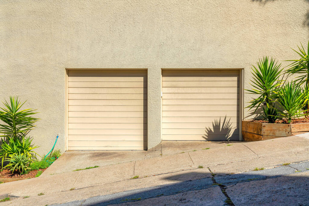 Две бежевые двери гаража в Сан-Франциско, Калифорния. Гараж снаружи с наклонной бетонной дорожкой и улицей с растениями сбоку дверей. - Фото, изображение