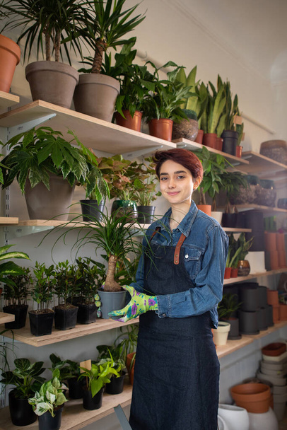 Smiling young entrepreneur in her nursery working in the greenhouse tending to the potted plants for sale with a glowing sun backdrop - Фото, зображення