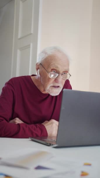 un homme âgé dans une grande pièce est assis à une table avec un ordinateur portable. Il utilise un casque sans fil pour écouter son interlocuteur via un appel vidéo - Séquence, vidéo