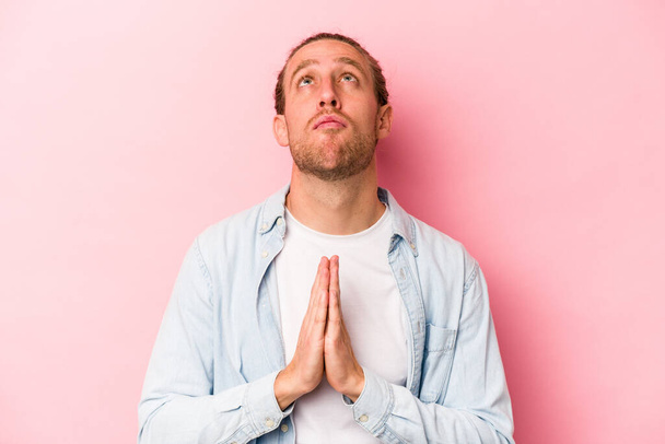 Young caucasian man isolated on pink background holding hands in pray near mouth, feels confident. - Fotoğraf, Görsel