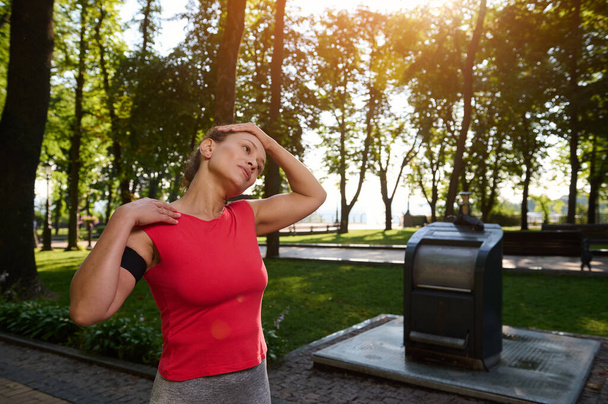 Latino urheilullinen nainen tiukka punainen t-paita älypuhelimen haltija seisoo kaupungin puistossa tekemässä venyttely liikuntaa, nauttia aamulla harjoitus tai lämmetä hänen ruumiinsa ennen aamulenkille - Valokuva, kuva