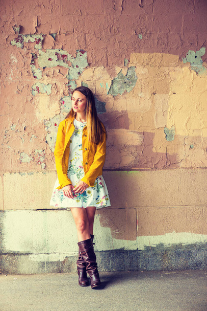 Woman Casual Fashion. A pretty  girl wearing flower patterned underdress, yellow corduroy jacket, brown leather high riding boots, standing by wall peeling off paints, looking away. - Photo, Image