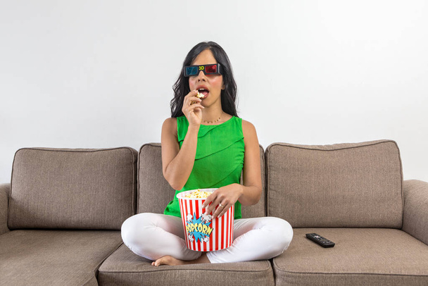 Spaanse vrouw in casual kleding en 3D-bril eten van verse popcorn en het kijken naar film op tv, terwijl zitten gekruiste benen op de bank in de dag thuis - Foto, afbeelding