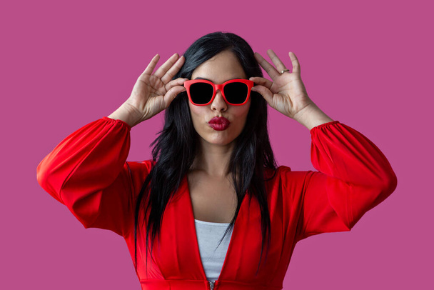 Trendy Hispanic woman in bright clothes pouting lips and adjusting sunglasses against purple background. - Photo, Image
