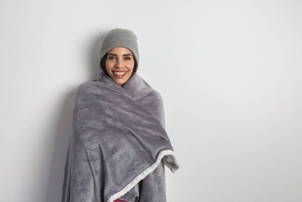 Feliz calentamiento femenino hispano con manta suave y gorro mientras está de pie sobre fondo blanco y mirando a la cámara - Foto, imagen