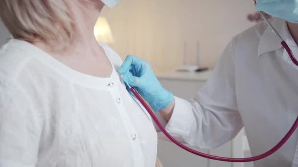 Una joven doctora con una bata blanca y una máscara usa un estetoscopio. Escucha los pulmones de una anciana guapa. Imágenes de alta calidad 4k - Imágenes, Vídeo