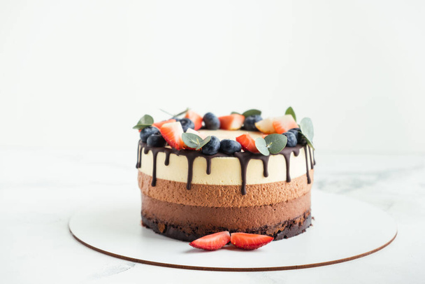 Κέικ με καφέ στρώσεις Τρία σοκολατάκια διακοσμημένα με bluberries, φράουλες και σταγόνες σοκολάτας στην κορυφή. Λευκό φόντο. Κοντινό πλάνο - Φωτογραφία, εικόνα