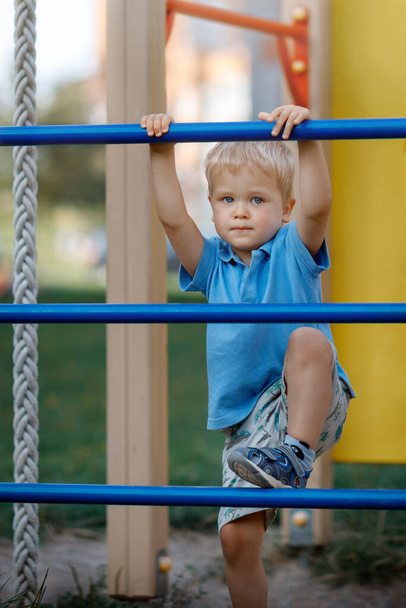 Small boy is climbing on a blue playground ladder. Looking at camera. Active child. Modern playground - Φωτογραφία, εικόνα