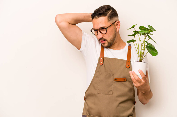 Ισπανόφωνος κηπουρός κρατώντας ένα φυτό απομονωμένο σε λευκό φόντο αγγίζοντας το πίσω μέρος του κεφαλιού, σκεπτόμενος και κάνοντας μια επιλογή. - Φωτογραφία, εικόνα