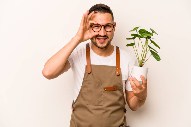 Hispanic gardener man holding a plant isolated on white background excited keeping ok gesture on eye. - Photo, Image