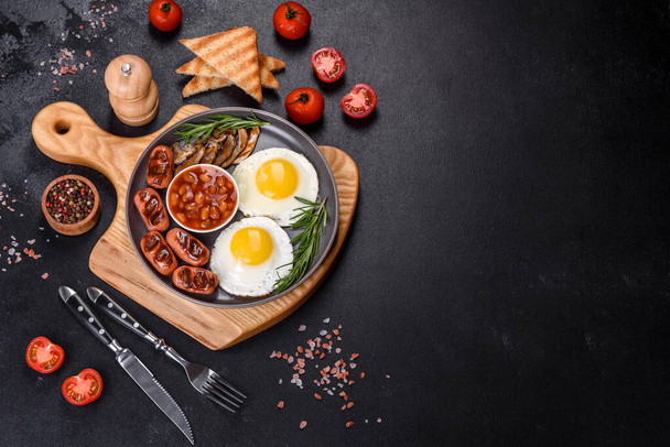 Πλήρες αγγλικό πρωινό - φασόλια, τηγανητά αυγά, ψητά λουκάνικα, ντομάτες, μανιτάρια σε σκουρόχρωμο τραπέζι με φρυγανισμένο ψωμί - Φωτογραφία, εικόνα