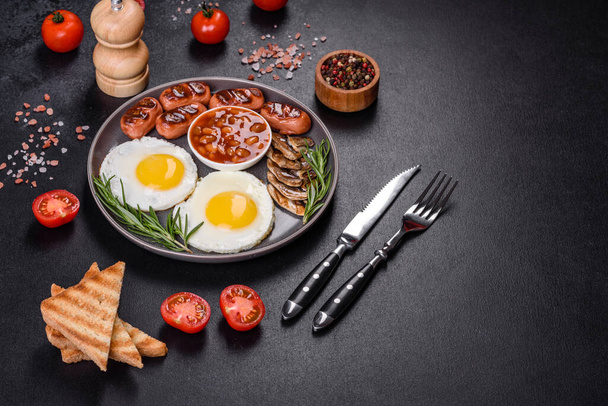 Plně anglická snídaně - fazole, smažená vejce, pečené klobásy, rajčata, houby na tmavém betonovém stole s opečeným chlebem - Fotografie, Obrázek
