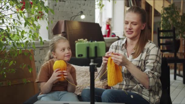 The girl and mother are engaged in needlework. The girl is holding a ball of wool, and the mother is knitting. In front of them is a phone on a tripod. They wave their hands at the camera and end the - Footage, Video