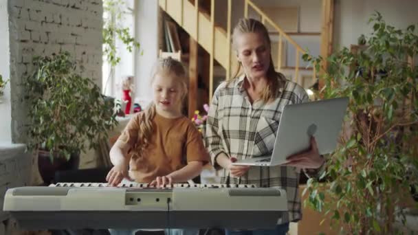 Мать помогает дочери тренироваться на синтезаторе. Она стоит рядом с ноутбуком и инструктирует ребёнка. Высококачественные 4k кадры - Кадры, видео