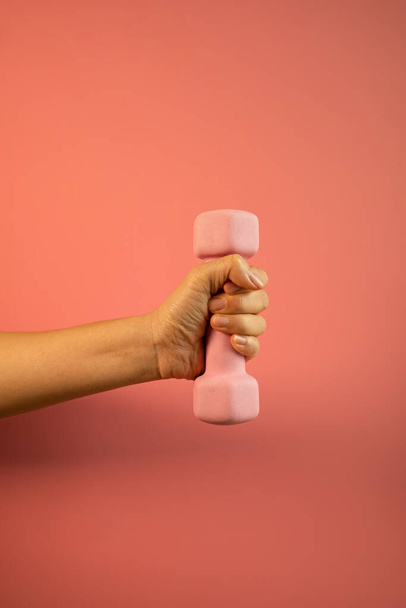 Egy nő keze egy rózsaszín súlyzót tart rózsaszín alapon elszigetelve. Felszerelés az otthoni edzéshez. Fitness és aktivitás. A sport és az egészséges életmód fogalma.  - Fotó, kép