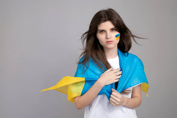 Mujer ucraniana de pie sobre un fondo gris aislado con bandera ucraniana azul y amarilla en su mejilla. el fondo de su diseño. Invasión rusa de Ucrania, Parar la guerra. - Foto, imagen