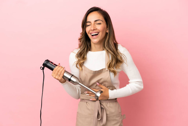 Young woman using hand blender over isolated pink background smiling a lot - Foto, afbeelding
