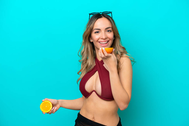 Young caucasian woman isolated on blue background in swimsuit and holding an orange - Foto, imagen