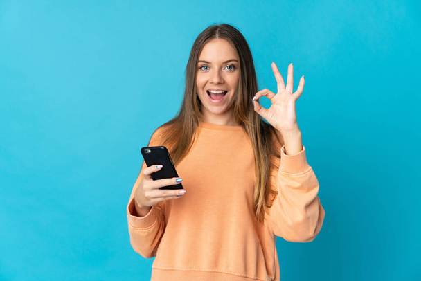 Young Lithuanian woman isolated on blue background using mobile phone and doing OK sign - Foto, imagen