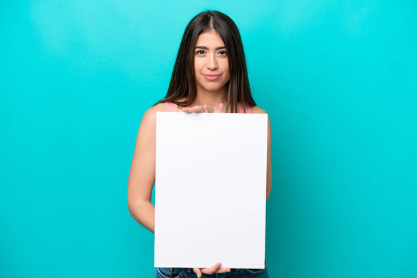 Молодая белая женщина, изолированная на синем фоне, держит пустой плакат со счастливым выражением лица - Фото, изображение
