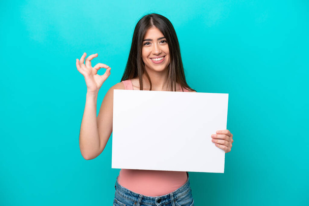 Mladá běloška izolované na modrém pozadí drží prázdný plakát a dělá OK znamení - Fotografie, Obrázek