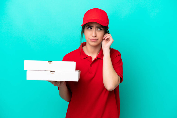 İş üniformalı bir pizzacı kadın mavi arka planda izole edilmiş pizza kutuları alıyor hayal kırıklığına uğramış ve kulaklarını kapatıyor. - Fotoğraf, Görsel