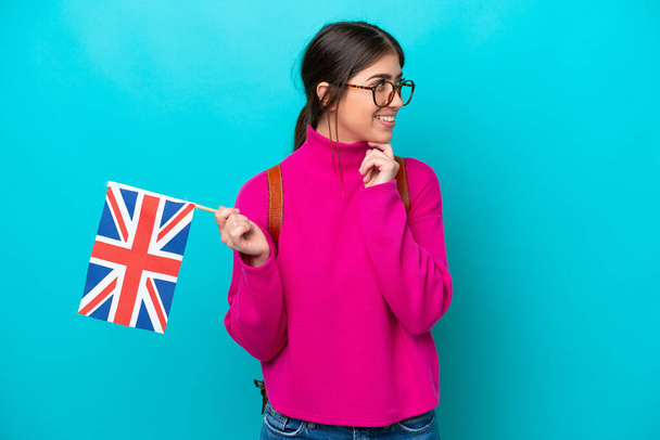 Young caucasian student woman holding English flag isolated on blue background looking to the side and smiling - Foto, imagen