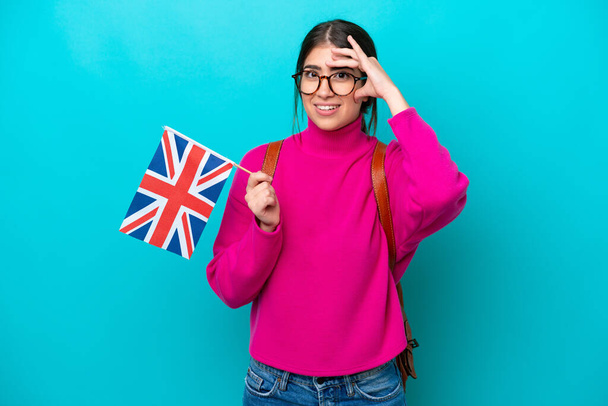 Young caucasian student woman holding English flag isolated on blue background doing surprise gesture while looking to the side - Photo, image