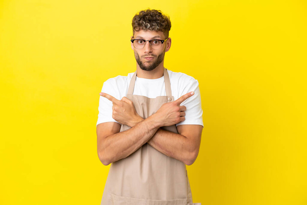 Restaurant waiter blonde man isolated on yellow background pointing to the laterals having doubts - Zdjęcie, obraz